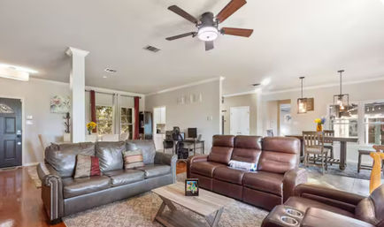 Bright open-concept living room with dark leather sofas and recliner, rustic wood coffee table, ceiling fan, hardwood floors and adjoining dining area with pendant lights and large windows.