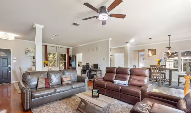 Bright open-concept living room with dark leather sofas and recliner, rustic wood coffee table, ceiling fan, hardwood floors and adjoining dining area with pendant lights and large windows.