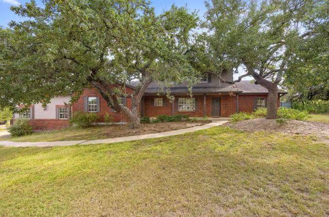 Red brick suburban home with covered front porch and dormer, shaded by sprawling oak trees with a curved concrete walkway and grassy front yard