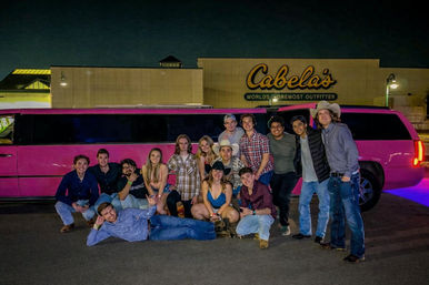 Young adults posing by a bright pink stretch limousine in a nighttime parking lot outside a large outdoor retailer storefront, some wearing casual and cowboy-style outfits, smiling for a group photo.