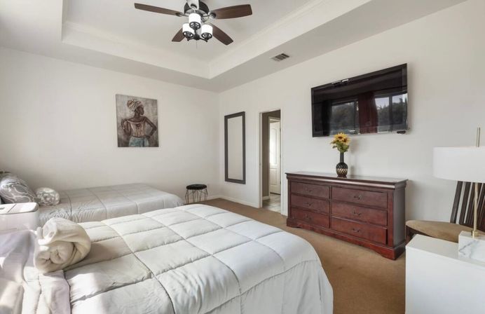 Bright neutral modern bedroom with two beds, tray ceiling and ceiling fan, wall-mounted flat-screen TV above a dark wooden dresser topped with a sunflower vase, white bedding and rolled towel
