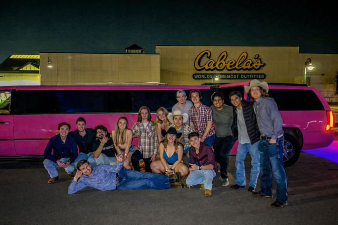 Group photo of friends in cowboy hats and jeans posing by a pink stretch limo outside an outdoor outfitter store at night in a parking lot.