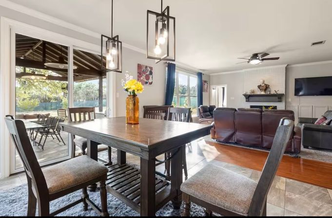 Sunlit open-concept dining and living room with a dark wood farmhouse table and upholstered chairs, amber vase with yellow flowers, black geometric pendant lights, sliding glass doors to a covered patio and backyard, leather sofas, fireplace, ceiling fan and wall-mounted TV.