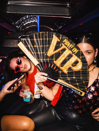 Two friends partying inside a night bus, one holding a black-and-gold folding fan reading "VIP", wearing red cat-eye sunglasses and sequin clubwear, phones and drinks in hand under colorful disco lights.