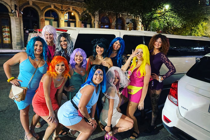 Smiling group of women in colorful wigs and party outfits posing by a white stretch limousine on a downtown city street at night for a bachelorette celebration.