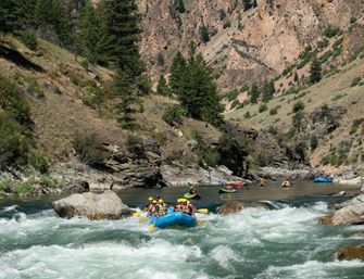 Whitewater rafting group paddling a blue inflatable through rapids on a rocky mountain river canyon with pine-covered slopes and distant kayakers — outdoor adventure scene