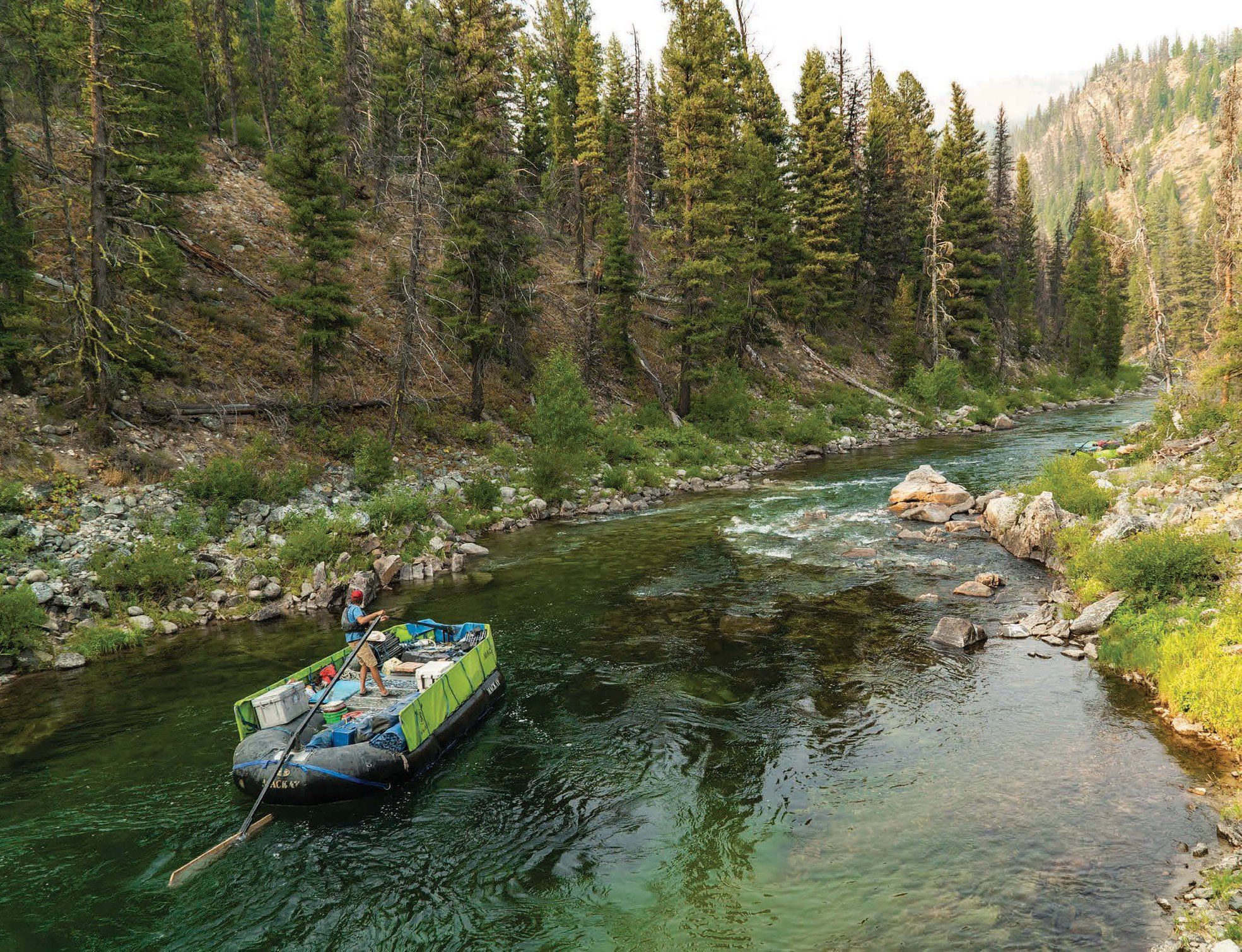 Solo rafter poling a loaded inflatable raft down a clear mountain river flanked by pine forest, rocky banks and distant peaks