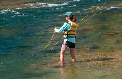 Person wading in a shallow river on a sunny day, wearing a life jacket and cap while casting a bright yellow fly-fishing line over clear, gently flowing water.
