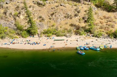 Aerial view of a sandy river beach with colorful inflatable rafts docked along green water, tents and camp gear lined beneath a rocky, tree-covered hillside, people wading and relaxing on a sunny day.
