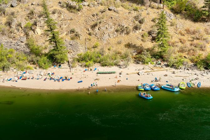 Aerial view of a sandy river beach with colorful inflatable rafts docked along green water, tents and camp gear lined beneath a rocky, tree-covered hillside, people wading and relaxing on a sunny day.