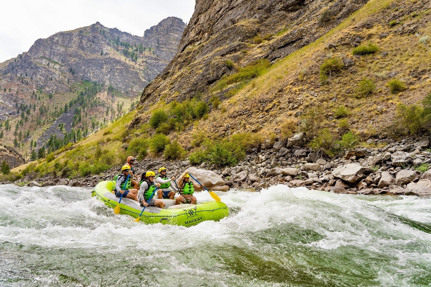 Group whitewater rafters in a bright inflatable raft paddling through rapids in a rocky mountain river canyon — outdoor adventure with helmets and life jackets