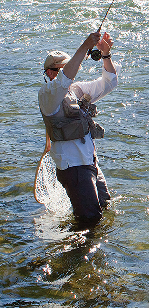 Waist-deep angler fly-fishing in a sunlit river, casting with a rod and reel while a mesh landing net trails in the water.