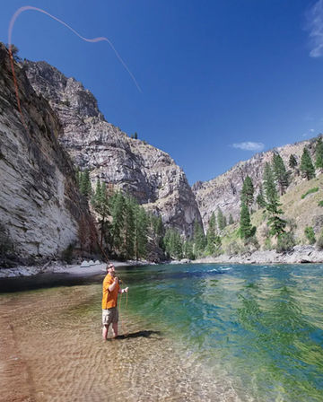 Angler in an orange shirt fly-fishing waist-deep in a clear green mountain river below steep rocky canyon walls and pine-covered slopes under a bright blue sky.