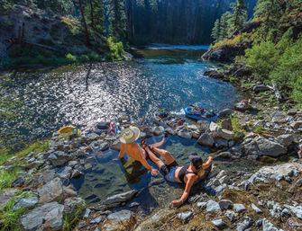 Two people soaking in a rocky riverside hot spring pool above a sparkling river with inflatable rafts on the shore and a pine-covered canyon beyond.