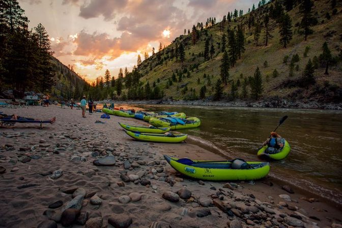 Green inflatable kayaks beached on a sandy, rocky riverbank at sunset, pine‑covered hills rising behind and campers preparing for a scenic mountain river float.