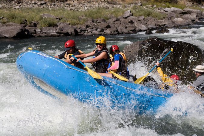 Whitewater rafting on a rocky mountain river: group in a bright blue inflatable raft paddling through splashing rapids, wearing helmets and life jackets as a guide steers.