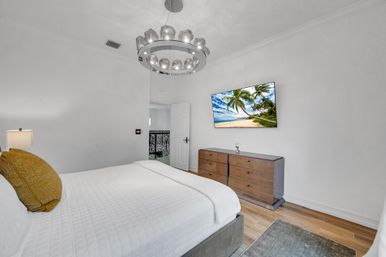 Bright modern bedroom with white walls and hardwood floors, white quilted bed with mustard pillow, circular crystal chandelier, wall-mounted TV showing a tropical beach above a wooden dresser, open door revealing ornate wrought-iron railing.