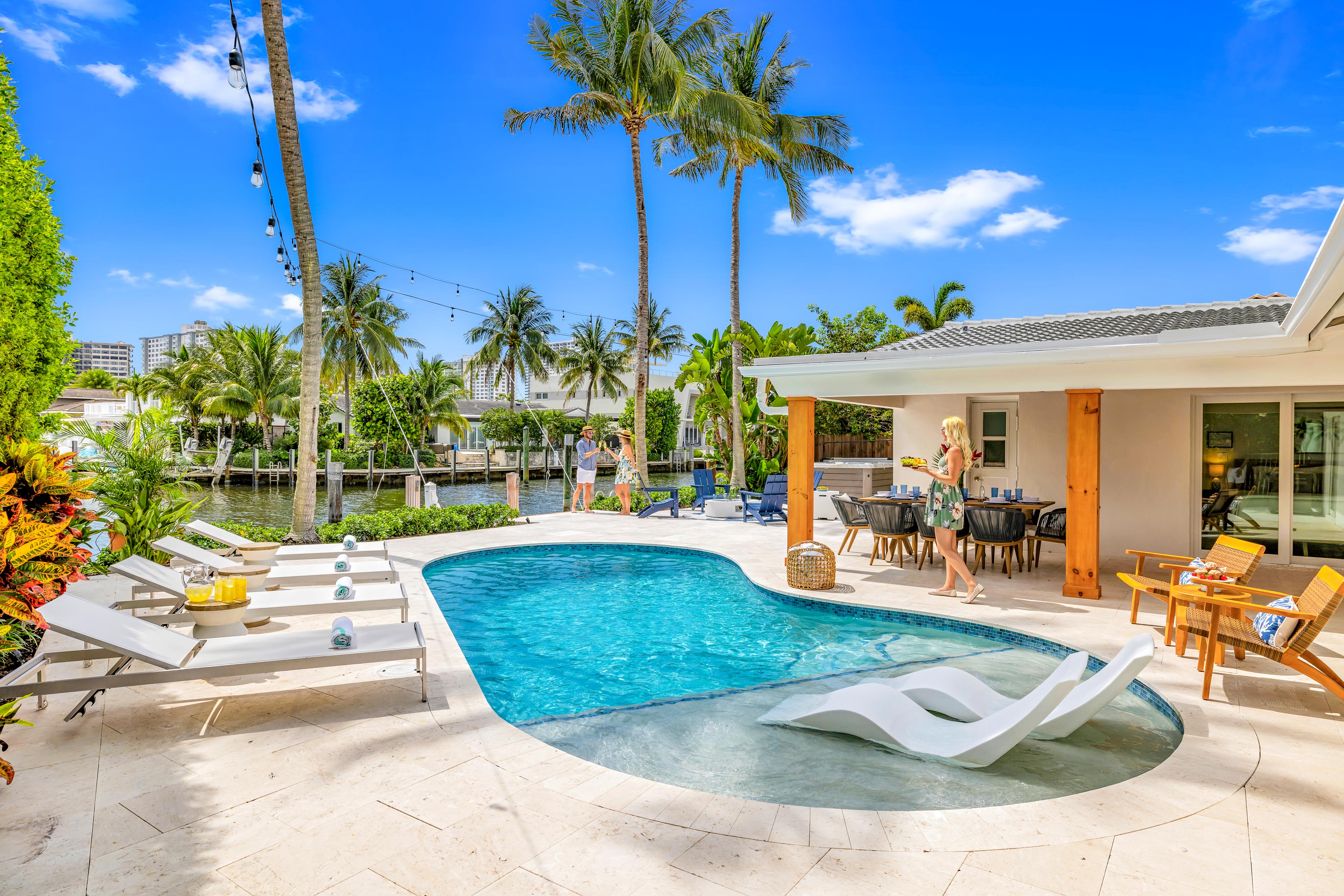 Sunny tropical waterfront backyard with palm trees and canal view, kidney-shaped pool with in-pool loungers, sunbeds, and covered patio dining area