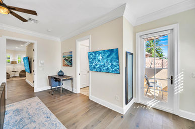 Sunny coastal-style home office with hardwood floors, small desk and blue swivel chair, wall-mounted TV displaying ocean imagery, full-length mirror, ceiling fan, area rug, and a glass door opening to a sunlit balcony with terracotta roof tiles and palm trees.