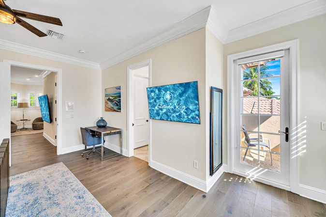 Sunny coastal-style home office with hardwood floors, small desk and blue swivel chair, wall-mounted TV displaying ocean imagery, full-length mirror, ceiling fan, area rug, and a glass door opening to a sunlit balcony with terracotta roof tiles and palm trees.