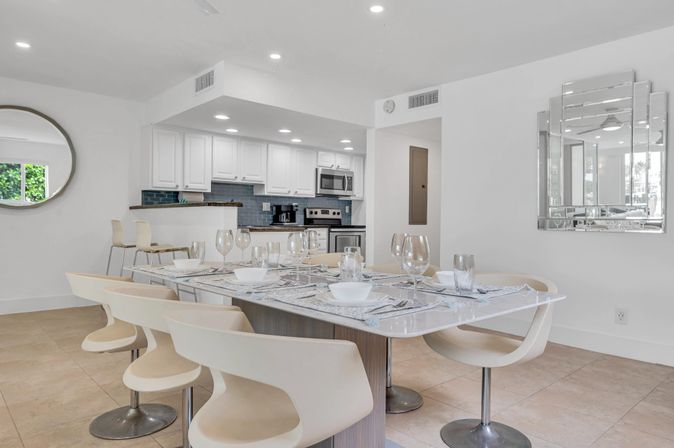 Bright open-plan dining area and modern kitchen — glass-topped table set for dinner with wine glasses, cream swivel chairs, white cabinets and stainless-steel appliances.