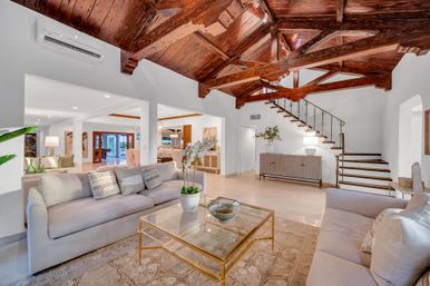 Open-concept living room with dramatic dark wood vaulted beams, neutral sofas, glass-and-gold coffee table, potted orchid, polished stone floors and a floating staircase.