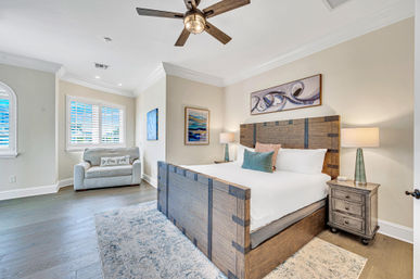 Bright coastal-style master bedroom with rustic wooden king bed, matching nightstands, shuttered windows, ceiling fan, and sleeper loveseat