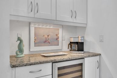 Coastal-inspired coffee station in a modern white kitchen: granite countertop, silver drip coffee maker, mint ceramic seahorse, glass-front wine fridge, and framed shorebird print lit by under-cabinet lights.