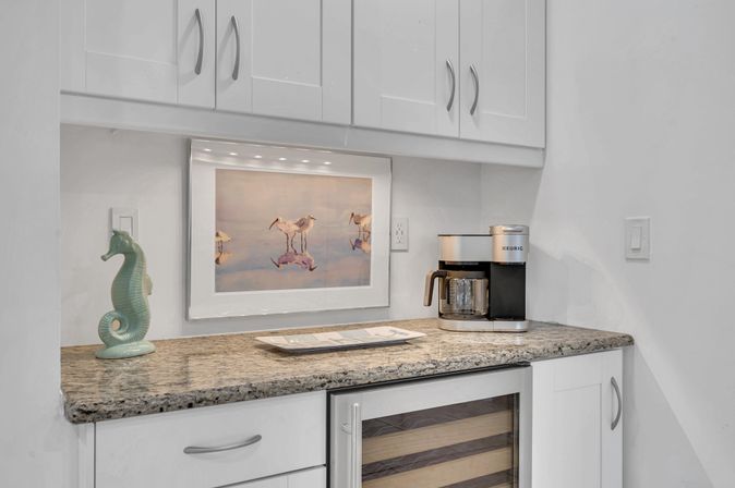 Coastal-inspired coffee station in a modern white kitchen: granite countertop, silver drip coffee maker, mint ceramic seahorse, glass-front wine fridge, and framed shorebird print lit by under-cabinet lights.