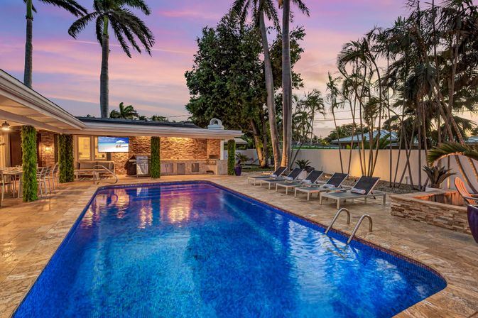 South Florida tropical backyard at sunset with an illuminated blue swimming pool, stone patio, outdoor kitchen and bar, row of lounge chairs and tall palm trees
