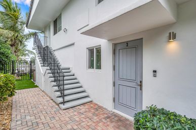 Cheerful coastal-style exterior showing unit 3 gray door, brick-paved walkway, black metal staircase to upper floor, white stucco walls, palm trees and tropical landscaping, modern wall sconce and doorbell.