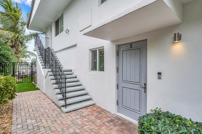 Cheerful coastal-style exterior showing unit 3 gray door, brick-paved walkway, black metal staircase to upper floor, white stucco walls, palm trees and tropical landscaping, modern wall sconce and doorbell.