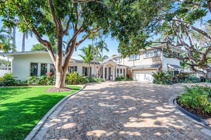 Sunlit two-story coastal-style home with brick paver circular driveway, large shade trees, palm trees and lush tropical landscaping.