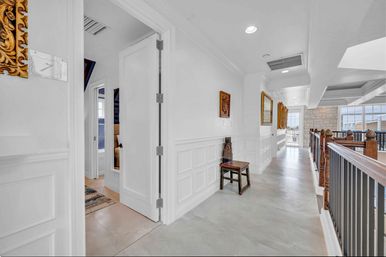 Bright second-floor hallway in a luxury home with white paneled walls, an antique wooden chair, framed art, wood-and-iron railing and large windows overlooking rooftops.