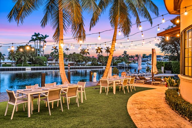 Tropical waterfront patio at sunset with string lights, palm trees, outdoor dining tables, lounge chairs and boats along a calm canal