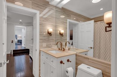 Bright contemporary bathroom with wood-look tile walls, white beadboard vanity, beige countertop and brass faucets, large mirror and wall sconces, toilet, shower curtain, and view into a bedroom with dark hardwood floors.