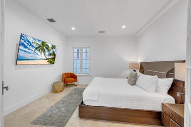 Bright modern minimalist bedroom with king bed and wood headboard, crisp white linens, wall-mounted TV showing a tropical beach scene, orange accent chair, geometric side table, gray runner rug and marble floor.