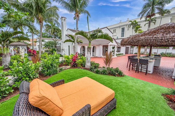 Wicker chaise lounge with orange cushion on vivid green lawn in a tropical courtyard with palm trees, colorful plants, a white Mediterranean-style villa and a thatched-roof tiki bar with outdoor kitchen and barstools on a red brick patio.