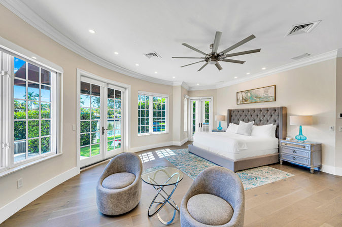 Sunlit waterfront master bedroom with king bed and tufted headboard, French doors and windows overlooking pool and garden, modern ceiling fan, hardwood floors and cozy seating area.