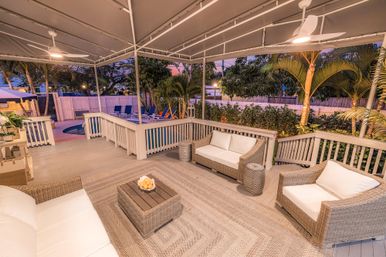 Cozy covered tropical backyard deck with wicker seating, woven rug, ceiling fans and a glowing pool at dusk