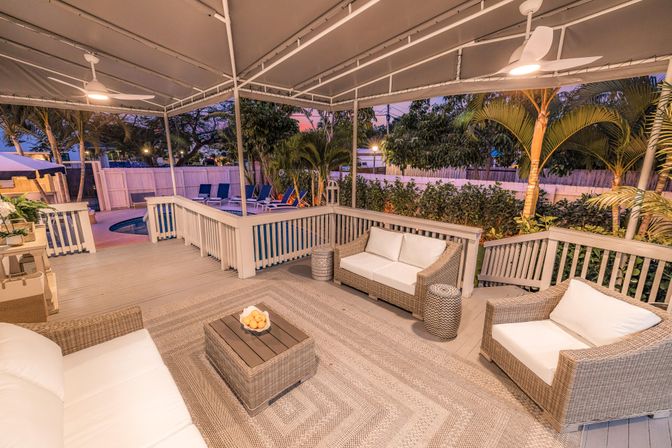 Cozy covered tropical backyard deck with wicker seating, woven rug, ceiling fans and a glowing pool at dusk
