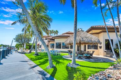 Waterfront, palm-lined tropical backyard at a Mediterranean-style villa with a thatched tiki hut patio, outdoor dining sets on vibrant green lawn and a wooden dock along the marina under a bright blue sky.