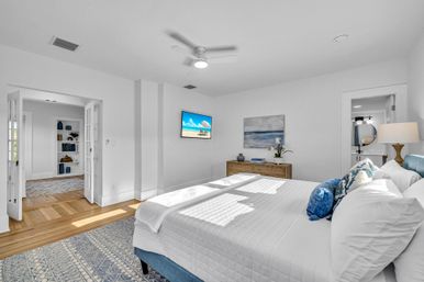 Sunlit coastal-style master bedroom with white quilted king bed, blue headboard and pillows, hardwood floors, patterned rug, wooden dresser with ocean art and wall-mounted TV, ceiling fan, and French doors to a hallway with built-in shelves and an en-suite bathroom.