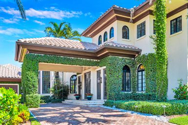 Sunny Mediterranean-style two-story home with terracotta tile roof, arched black-framed windows, ivy-covered portico, palm trees and brick driveway