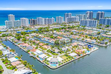 Aerial view of a sun-drenched coastal waterfront neighborhood with canal-lined residential islands, private boat docks, palm trees, and oceanfront high-rise condos under a bright blue sky.