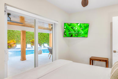 Sunny vacation bedroom with white bedding and wall-mounted TV, sliding glass doors opening to a covered patio and private blue pool with lounge chairs and tropical hedges