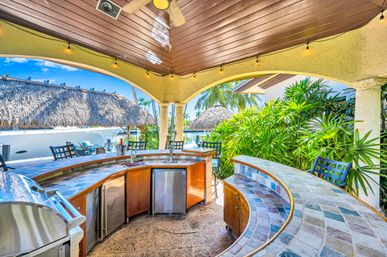 Sunny tropical waterfront outdoor bar under a wood-paneled roof with a curved tiled counter, built-in grill and stainless fridge, bar stools, ceiling fan, thatched palapa and lush palms
