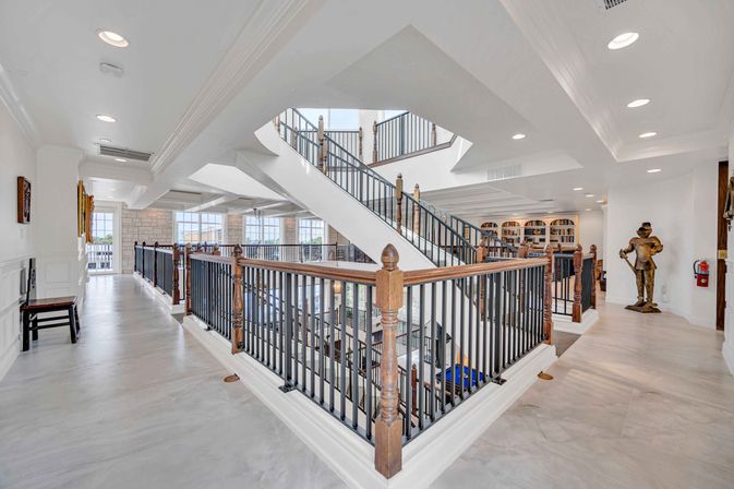 Sunlit luxury home atrium featuring a central white staircase with wood-and-iron railings, polished marble floors, floor-to-ceiling windows with water views, built-in bookshelves and a decorative knight statue.