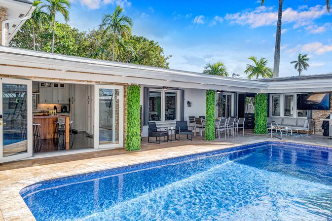 Sunny tropical backyard with blue-tiled pool, covered patio and outdoor dining, open sliding doors to a modern kitchen and palm trees.