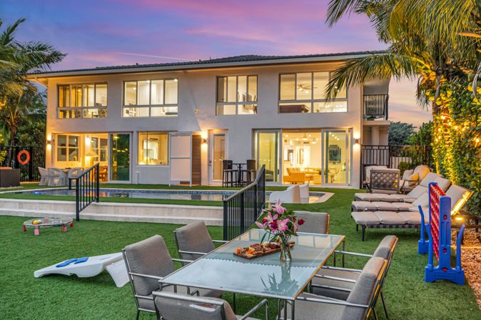 Dusk view of a luxury modern two-story home with glowing floor-to-ceiling windows, backyard pool, outdoor dining table with fresh flowers, lounge chairs, lawn games and palm trees in a tropical setting